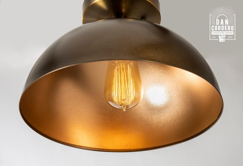 May include: A gold-toned metal dome pendant light fixture with a clear glass light bulb inside. The light is on and the bulb is glowing.