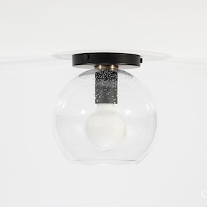 May include: A modern ceiling light fixture with a clear, speckled glass globe shade. The fixture has a black metal base and a white light bulb. The design is minimalist and contemporary, suitable for various interior styles.