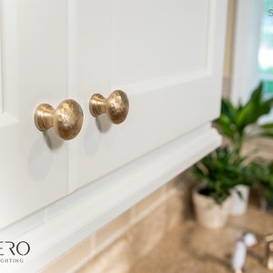 May include: Close-up of two satin brass finish cabinet knobs with a hammered texture. The knobs are mounted on a white cabinet door. The text "Satin Brass Finish" is visible in the upper right corner.