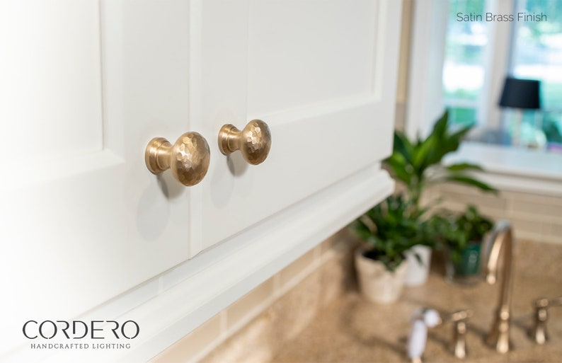 May include: Close-up of two satin brass finish cabinet knobs with a hammered texture. The knobs are mounted on a white cabinet door. The text "Satin Brass Finish" is visible in the upper right corner.