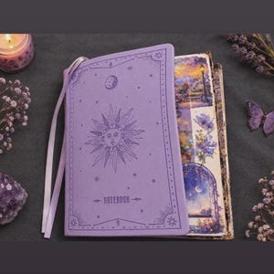 Könnte beinhalten: Ein hellviolettes Notizbuch mit einem geprägten Sonnen- und Mondmotiv auf dem Einband. Der Einband zeigt unten das Wort NOTEBOOK, dekorative Eckverzierungen und ein passendes Lesezeichenband. Die offenen Seiten zeigen eine bunte Blumenillustration.