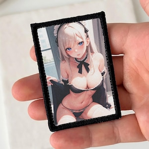 Pode incluir: Um emblema retangular bordado com uma personagem de estilo anime estampada, com longos cabelos loiros, olhos azuis e uma faixa preta. A personagem veste um conjunto preto e branco com um laço no pescoço, lingerie preta e meias brancas.