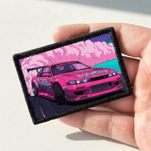 Peut inclure: Un écusson rectangulaire brodé représentant une voiture de sport rose vif en train de déraper sur une route sombre. La voiture est entourée de nuages de fumée rose stylisés et porte des inscriptions bleues style graffiti sur le capot et les côtés.