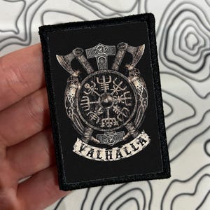 May include: A rectangular black patch featuring a detailed graphic in shades of silver and white. The design includes two crossed axes, a central circular shield with runic symbols, and a banner at the bottom with the text VALHALLA printed.