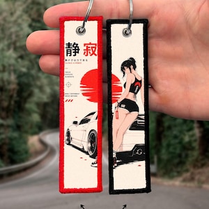 Peut inclure: Deux porte-clés rectangulaires brodés avec des anneaux métalliques. L'un a une bordure rouge, une voiture blanche, un soleil rouge et le texte japonais 静寂. L'autre a une bordure noire, un graphique d'une personne appuyée contre une voiture et un soleil rouge.
