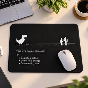 May include: A rectangular black mouse pad featuring a white pixelated dinosaur and cactus graphic. The text reads: There is no Internet connection. Try: Go make a coffee, Go out for a change, Do something else. A white computer mouse rests on the pad.