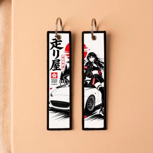 May include: Two rectangular fabric keychains with black stitched borders and metal rings. One features Japanese text, a red sun, and a white car on a mountain road. The other displays an anime-style character sitting on a white car with a red sun background.