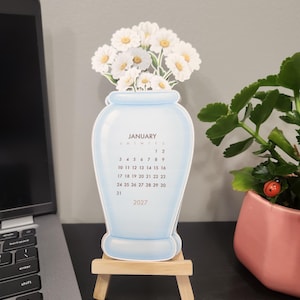 May include: A die-cut paper calendar for January 2027 shaped like a light blue vase filled with white daisies. The calendar is displayed on a small wooden easel stand, featuring the month name, days of the week, and dates printed in black.