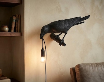 May include: A black wall-mounted lamp shaped like a crow perched on a branch. The crow holds a black electrical cord in its beak, which suspends a single glowing light bulb downward. The fixture is matte black with detailed feather textures.