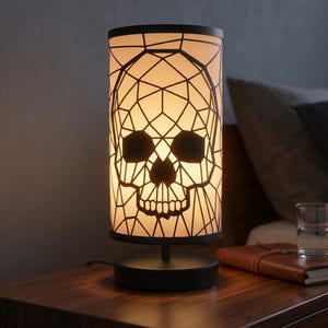 May include: A cylindrical table lamp featuring a black geometric frame that forms a skull design. The lamp has a warm, illuminated white shade and a solid black circular base, providing a soft glow through the intricate cutout pattern.
