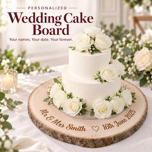 May include: A round wooden cake board with natural bark edges, engraved with the text 'Mr & Mrs Smith', a heart symbol, and '16th June 2025'. A two-tiered white frosted cake decorated with white roses and greenery sits on the wooden board.