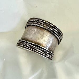 May include: A wide silver band ring featuring a smooth, polished center section flanked by two decorative borders with a raised, braided rope pattern. The metal has a slightly tarnished finish, highlighting the intricate texture of the edges.