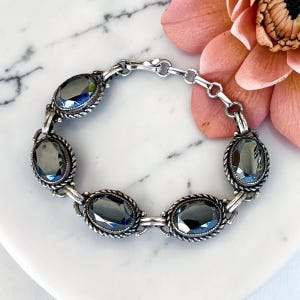 May include: A silver-toned link bracelet featuring five oval-shaped, dark gray faceted stones. Each stone is set within a twisted rope-style metal frame. The bracelet includes a secure metal clasp and a series of small circular connecting links.