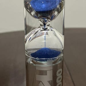 May include: A clear glass hourglass filled with vibrant blue sand. The hourglass features a narrow neck and a cylindrical base with the white printed text "EXL 2000" visible on the lower section of the glass container.