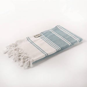 May include: A folded white and blue beach towel with a striped and diamond pattern. The towel has a fringe on one end and a small tag that reads "AKSEAN". The towel is made of a soft, absorbent material.