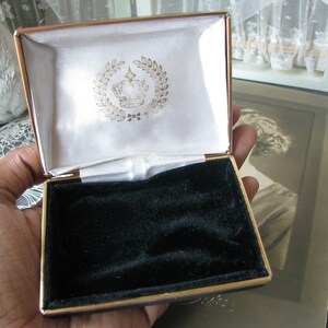 May include: A gold-colored jewelry box with a black velvet interior. The lid is open and features a white satin lining with a gold crown and laurel wreath design.