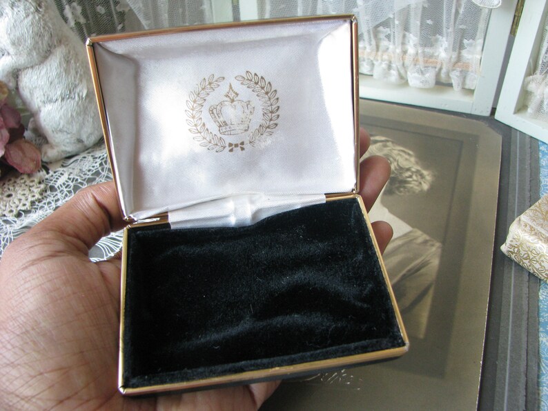May include: A gold-colored jewelry box with a black velvet interior. The lid is open and features a white satin lining with a gold crown and laurel wreath design.