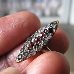 Puede incluir: Un anillo de oro con un dise&ntilde;o ovalado puntiagudo engastado con peque&ntilde;as piedras preciosas transparentes y rojas.