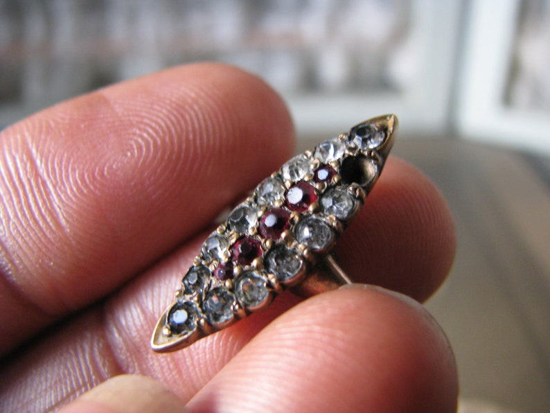 Puede incluir: Un anillo de oro con un dise&ntilde;o ovalado puntiagudo engastado con peque&ntilde;as piedras preciosas transparentes y rojas.