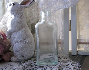 Puede incluir: Una botella de vidrio transparente con forma cuadrada y parte superior redondeada. La botella está sobre un mantel de encaje blanco.