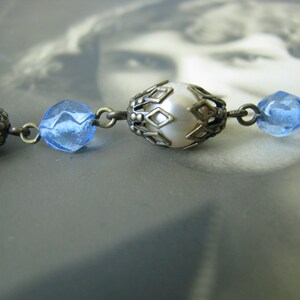 May include: A close-up of a vintage necklace with alternating white pearl and blue glass beads. The beads are held in place by ornate metal settings.