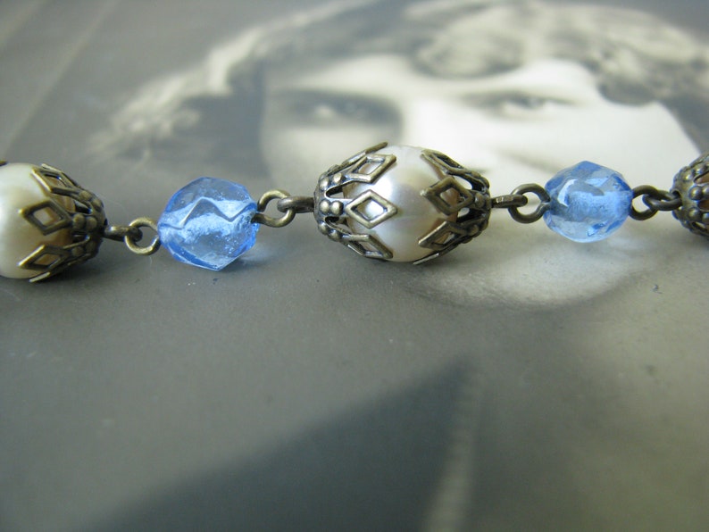 May include: A close-up of a vintage necklace with alternating white pearl and blue glass beads. The beads are held in place by ornate metal settings.