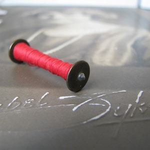May include: A small spool of red thread with a metal cap on each end. The spool is lying on a gray surface with white writing.