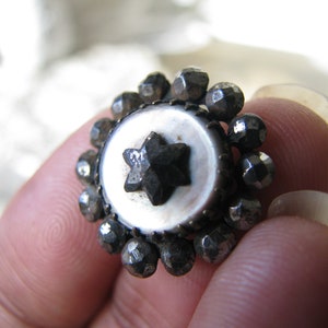 Puede incluir: Un botón de plata con un centro de perla blanca y una estrella negra en el centro. El botón está rodeado por un círculo de pequeñas cuentas de plata.