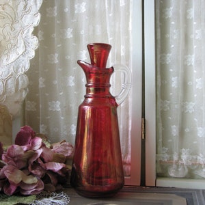 May include: A vintage ruby red glass bottle with a clear glass handle and stopper. The bottle is sitting on a wooden surface in front of a white lace curtain.