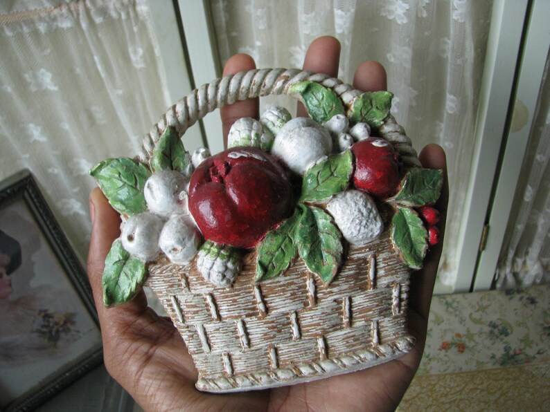 May include: A white ceramic basket filled with red and white fruit, with green leaves. The basket is hand-painted and has a rustic look.