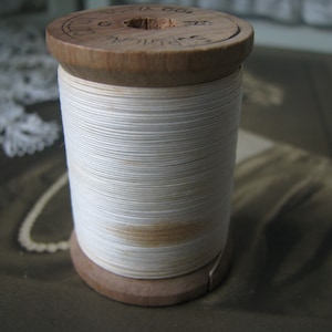 Op de afbeelding: Een vintage houten spoel met wit garen. De spoel heeft een lichtbruine houten basis en bovenkant, met het garen strak om het midden gewikkeld. De bovenkant van de spoel heeft gestempelde tekst. Het garen heeft een crèmewitte kleur.