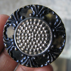 Puede incluir: Un botón redondo negro con un centro plateado. El centro está cubierto con un patrón texturizado en forma de diamante. El botón tiene un diseño floral decorativo alrededor del borde.