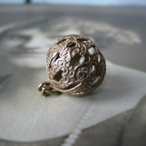 May include: A detailed, openwork metal sphere with a floral design. The sphere is a warm gold color and has a small chain attached. The intricate pattern and vintage aesthetic suggest it may be a pendant or charm.