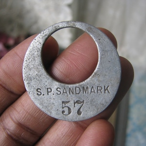 May include: A silver metal tag with the text "S.P. SANDMARK 57" engraved on it.