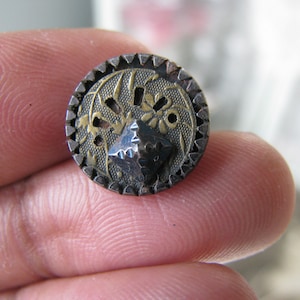May include: A round, silver button with a gold-colored design and a raised, pointed center. The button has a decorative, scalloped edge.