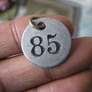 May include: A silver metal tag with a black number 85 on it. The tag has a small brass loop at the top.