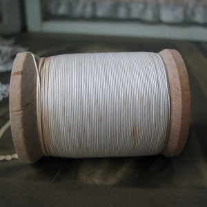 Op de afbeelding: Een spoel wit garen gewikkeld om een houten spoel. Het garen is glad en heeft een lichte glans.