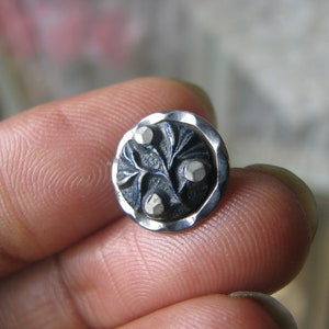 Puede incluir: Un botón plateado con un centro negro que presenta un diseño tallado de un árbol con tres hojas. Las hojas tienen acentos plateados en relieve.