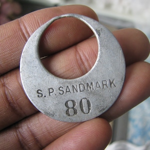 May include: A silver metal tag with the text "S.P. SANDMARK 80" engraved on it. The tag has a hole in the top and is shaped like a crescent moon.