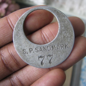 May include: A silver metal token with the text "S.P. SANDMARK 77" engraved on it.