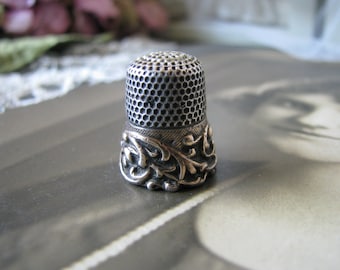 May include: A silver thimble with a decorative floral design around the base. The thimble has a textured top.
