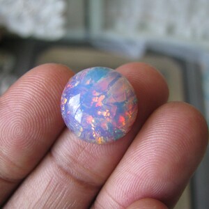 Può includere: Un cabochon di opale rotondo e iridescente con uno schema di colori blu chiaro, rosa e arancio. L'opale è incastonato in una montatura a bezel e viene tenuto in una mano.