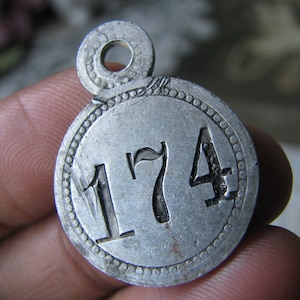 May include: A silver metal tag with a circular shape and a hole at the top. The tag has the number "174" engraved on it.