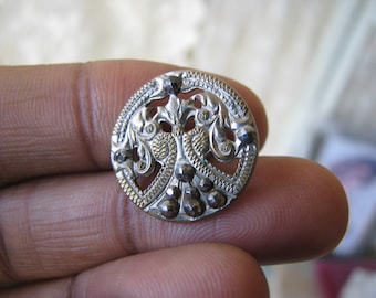 Op de afbeelding: Een ronde zilveren en gouden knoop met een decoratief ontwerp van gestileerde bloemen en bladeren. De knoop heeft een verhoogde rand en is versierd met kleine, ronde, zwarte kralen.
