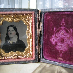 May include: A vintage, black, hinged, velvet-lined photo case with a gold-trimmed, ornate frame containing a black and white portrait of a woman with a white collar.
