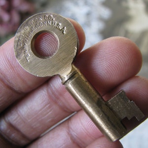 Könnte beinhalten: Nahaufnahme eines antiken Messingschlüssels. Der Schlüssel hat einen kreisförmigen Kopf mit dem Text "EAGLE LOCK CO. TERRYVILLE, CONN. U.S.A." Schaft und Bart des Schlüssels sind ebenfalls sichtbar. Der Schlüssel wird in einer Hand gehalten.