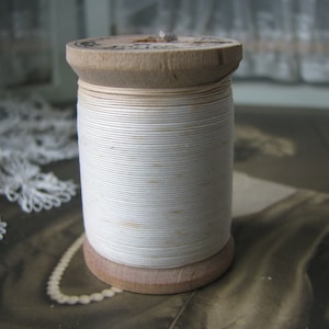 Op de afbeelding: Een houten spoel met witte draad. De spoel is gewikkeld met witte draad en heeft een houten boven- en onderkant.