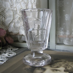 May include: A clear glass shot glass with a decorative, geometric pattern. The glass has a wide base and a short, straight stem.
