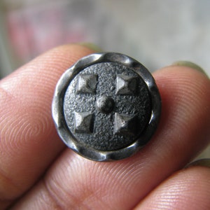 Puede incluir: Un botón de metal plateado con un diseño texturizado en relieve. El botón tiene forma circular con un borde elevado y cinco acentos en forma de estrella de cinco puntas.