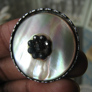 Puede incluir: Un botón de plata y nácar con un grupo de piedras preciosas negras en el centro.